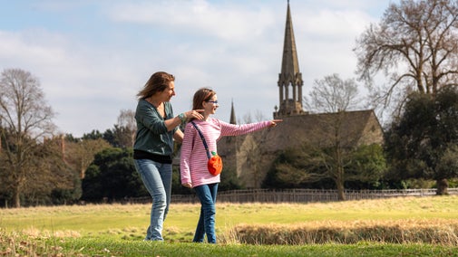 A mother and daughter explore Charlecote's parkland on a sunny day with St Leonard's church in the background